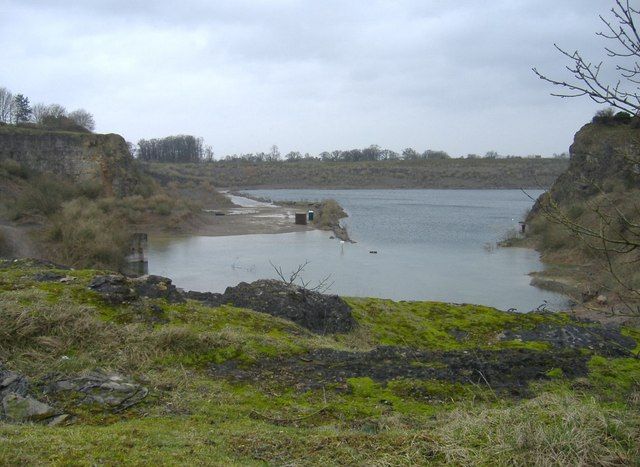 A view of Cromhall Quarry from the south side on an overcast day. Sheer, layered rock cliffs surround the large, calm body of deep blue water. Green vegetation and mosses grow on the upper edges of the cliffs and in the foreground.